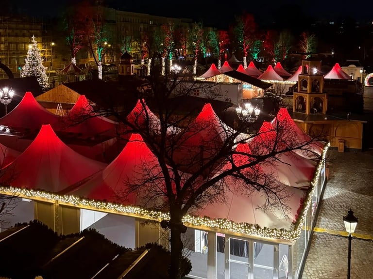 Nighttime holiday market with red tent canopies, bare tree, and golden light garland decorations along storefronts