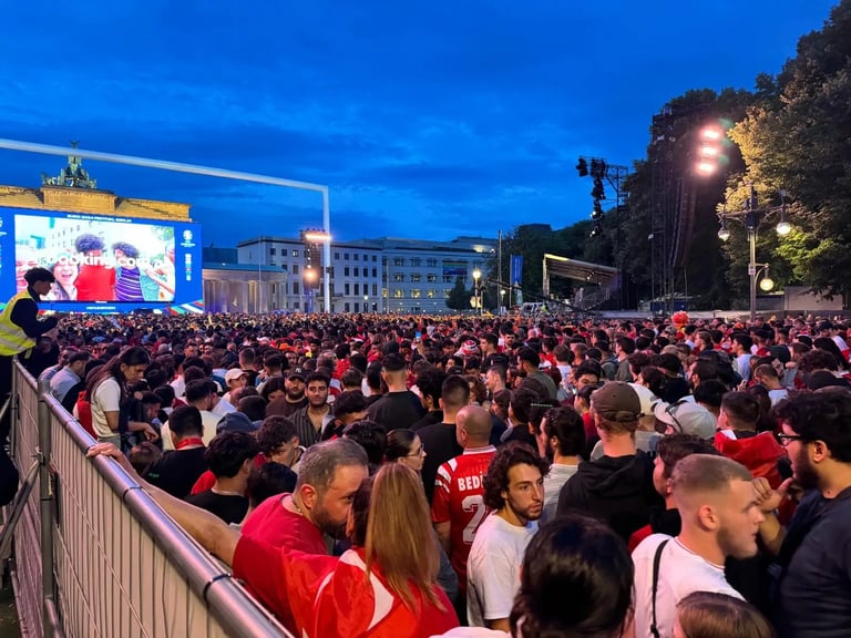 Large crowd of people gathered at an outdoor public square at dusk, watching a large screen display, with buildings and street lights visible in the background.